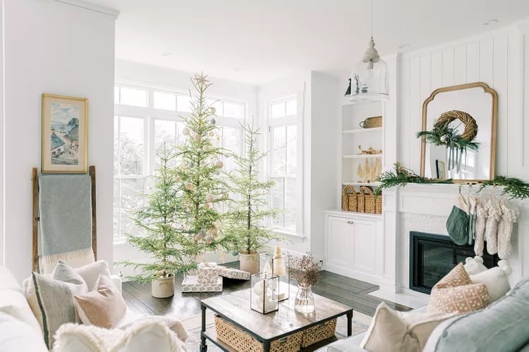 Living room decorated with 3 christmas trees, white chairs, white built-in shelving, and stockings on the fireplace mantel from our holiday decor updates guide.