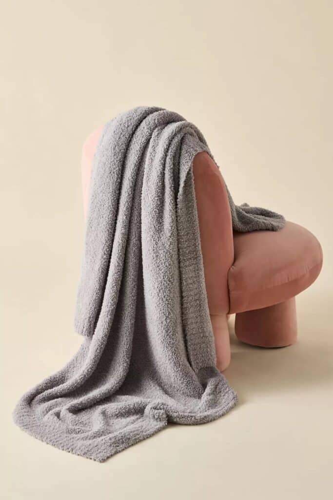 Urban Outfitters Barefoot Dreams Throw Blanket in Dove Gray is draped over a pink velvet chair from our home decor gift guide.