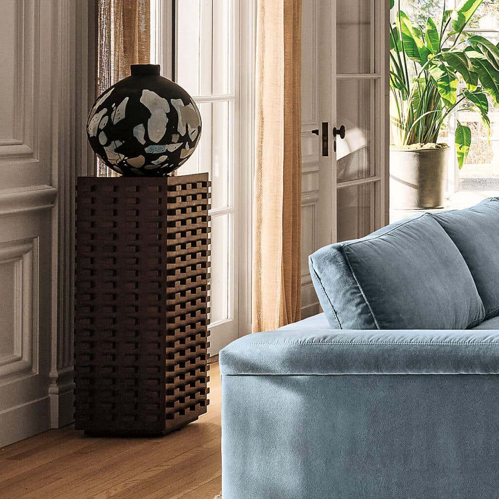 Living room with tall pedestal table supporting a black and silver vase behind a blue velvet sofa.
