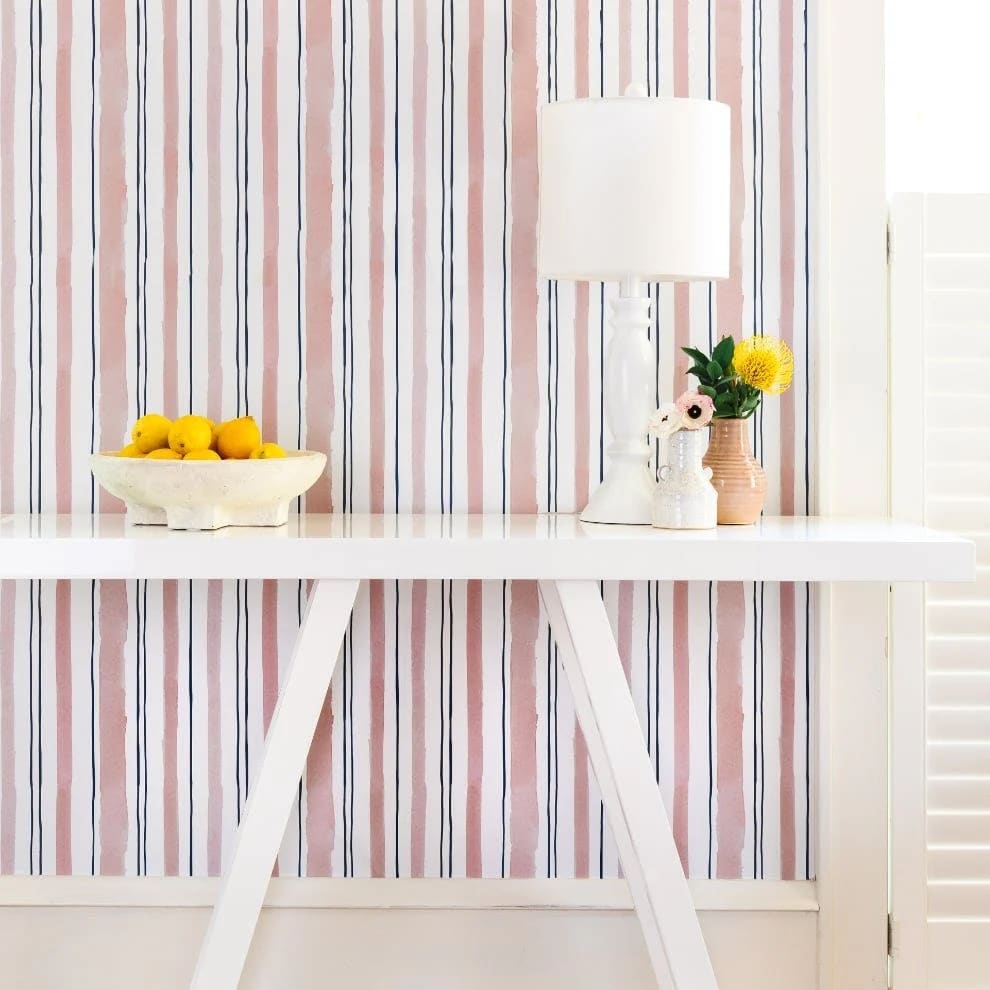 Обои Chasing Paper с полосами в темно-синем и розовом цветах за белой консолью с белой лампой и белой миской с лимонами.